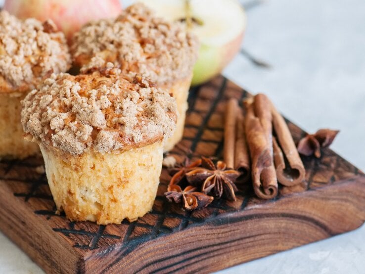 Drei Apfel-Streusel-Muffins auf einem Brett mit Anissternen und Zimtstangen, im Hintergrund unscharf halbierte Äpfel.