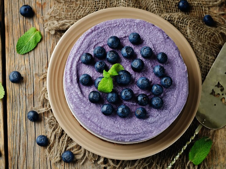 Auf einem Holztisch steht ein Heidelbeer-Käsekuchen ohne Backen. Er ist mit Blaubeeren dekoriert. die Creme ist lila eingefärbt.