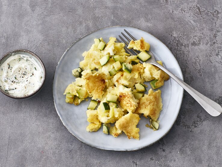 Ein heller Teller mit Halloumi-Zucchini-Schmarrn und einer Gabel steht auf einem grauen Untergrund. Links neben dem Teller steht eine kleine Schmale mit Schmarrn-Dip.