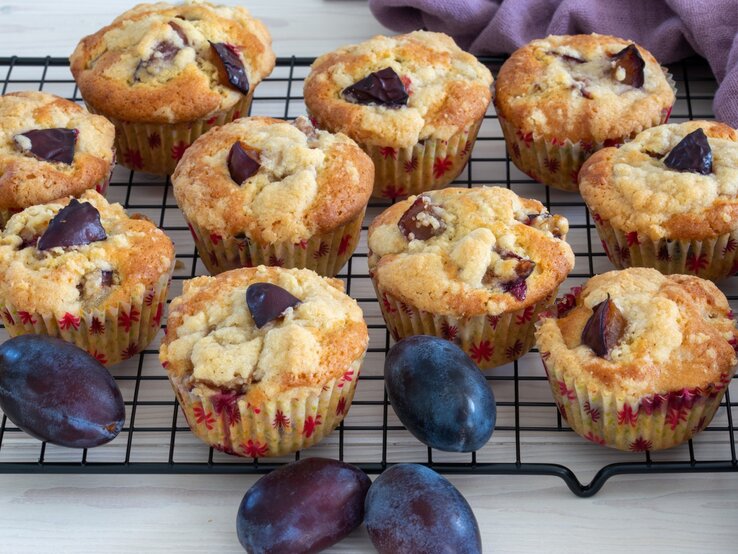 Pflaumen-Muffins mit griechischem Joghurt liegen auf einem Gitter. Daneben liegt ein violettes Tuch und einige Pflaumen. 