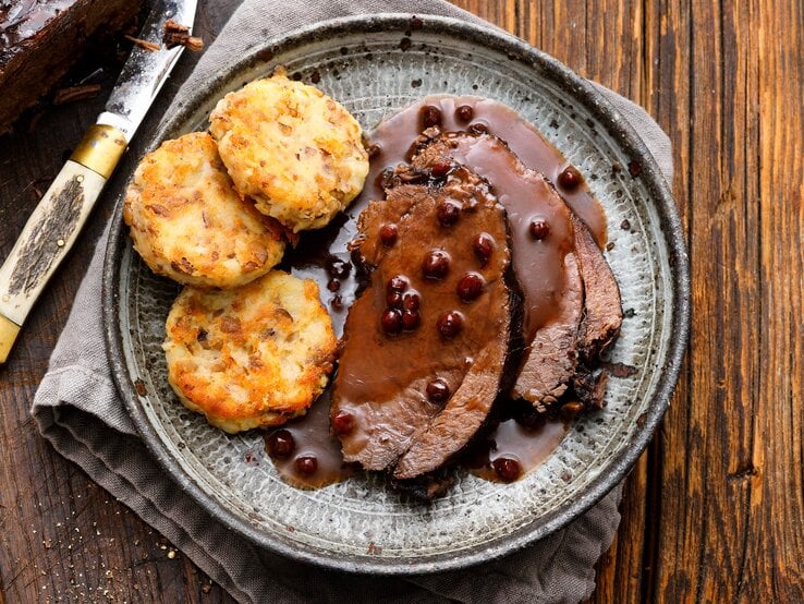 Ein Teller auf dem der Wilde Sauerbraten in Lebkuchensoße mit Kartoffeln-Maronen-Münzen angerichtet ist, neben einem Messer, dem restlichen, angeschnittenen Braten, auf einem Tuch vor einem Holzhintergrund, von oben. 