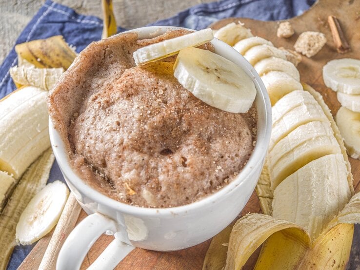 In einer Tasse ist ein Bananen-Tassenkuchen gebacken. Oben drauf liegen zwei Scheiben Bananen. Er ist mit Zimt bestreut.