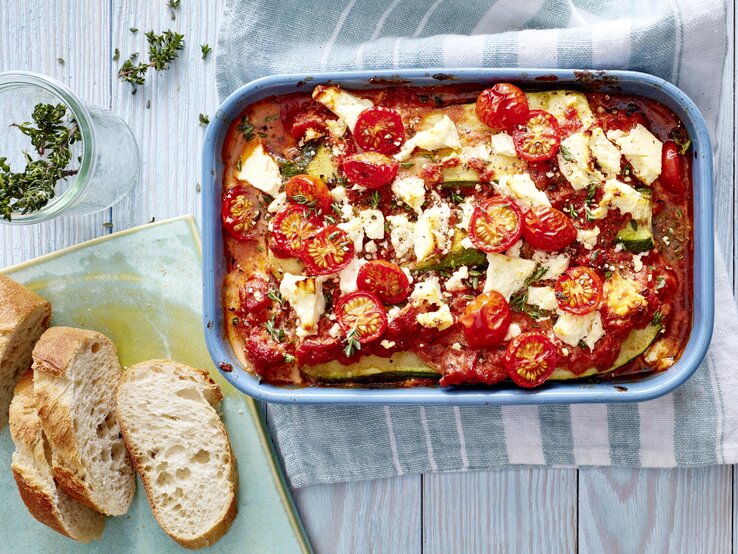 Eine Zucchini-Lasagne mit Feta in einer blauen Auflaufform steht auf einem Geschirrtuch auf einem blauen Tisch. Die Lasagne ist angerichtet mit Feta, Tomaten und Thymian. Neben der Auflaufform liegt Baguette und ein Glas mit Kräutern.