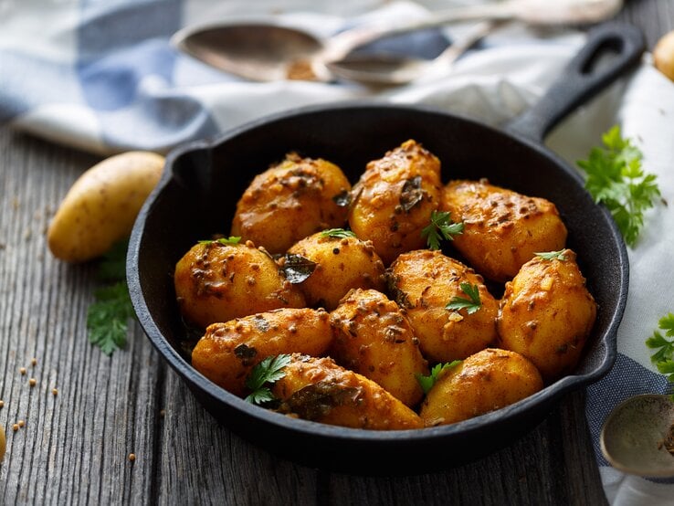 Eine gusseiserne Pfanne mit Bombay-Kartoffeln, bestreut mit frischen Kräutern, steht auf einem Holztisch. Drumherum sieht man ein Geschirrtuch, Besteck und einige Zutaten. | © Getty Images/ asab974