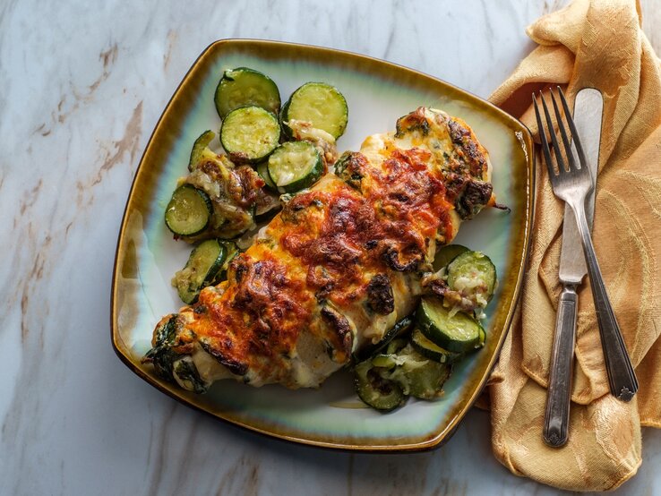 Hasselback Hähnchen mit Zucchini und Spinat auf einem farbigen Teller. Daneben Besteck auf einem Tuch. 