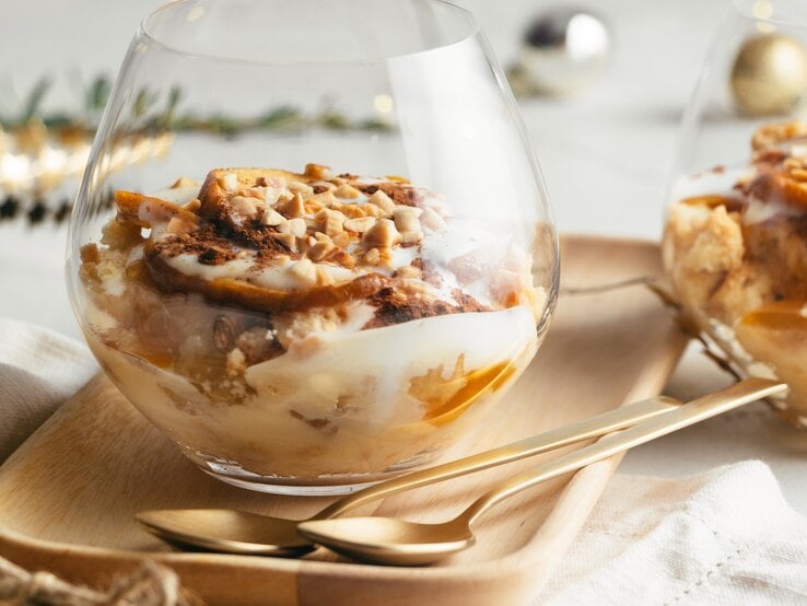 Bratapfel-Dessert im Glas mit Vanillesoße, Gewürzkaramell, Keksen und Mandelsplittern auf Holzteller, beiger Stoffserviette und Tisch mit weißem Tischtuch. Ein zweites Glas im Hintergrund angedeutet. Weihnachtdekoration in grün, gold, silber und Schwarz im Hintergrund unscharf. Aufnahme aus der Frontale.