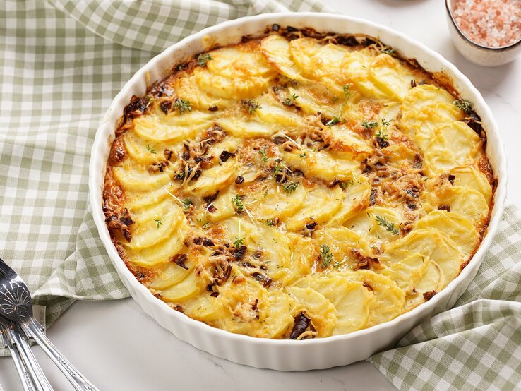 Eine runde weiße Auflaufform mit dem Kartoffelgratin auf einem weißen Tisch mit grün-weiß-karierter Tischdecke.