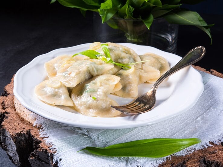 Frontal: Ein weißer Porzellanteller mit Bärlauch-Maultaschen steht auf einem weißen Tuch, das auf einem dunklen Untergrund liegt. Im Vorder- und Hintergrund sind frische Bärlauch-Blätter.