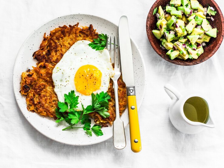 Ein weißer Teller mit den Röstis und Spiegelei auf einem weißen Tisch. Die Avocado steht daneben in einer kleinen braunen Schale.