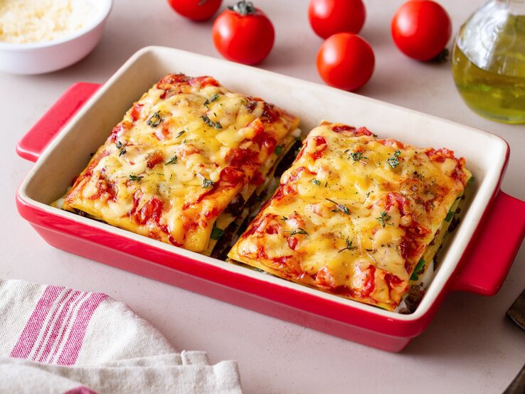 Bärlauch-Lasagne in roter Auflaufform mit Tomaten auf hellen Tisch. Rechts Flasche mit Olivenöl, links Schale mit Parmesan und Cocktailtomaten.