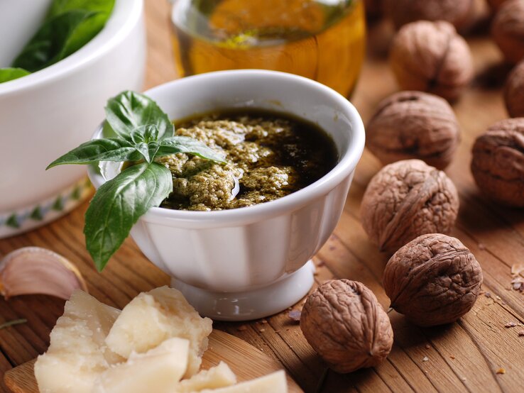 Frontalsicht: Auf einem Untergrund aus Holz steht ein weißes Schälchen selbst gemachtes Walnuss-Pesto mit einem Basilikumblatt als Deko. Drumherum liegen frische Walnüsse, geriebener Parmesan. Im Hintergrund ist eine Glasflasche mit Olivenöl.