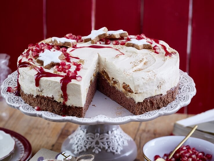 Eine angeschnittene Zimtstern-Tiramisu-Torte  mit Zimtsternen und Granatapfelkernen garniert, auf einem Kuchenständer neben einer Schüssel Granatapfelkerne vor einem roten Holzhintergrund. 