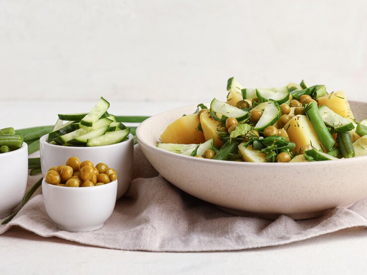 Eine weiße Schale mit dem Kartoffelsalat. Daneben drei kleine Schalen mit den weiteren Zutaten. Alles auf hellem Unter- und Hintergrund.