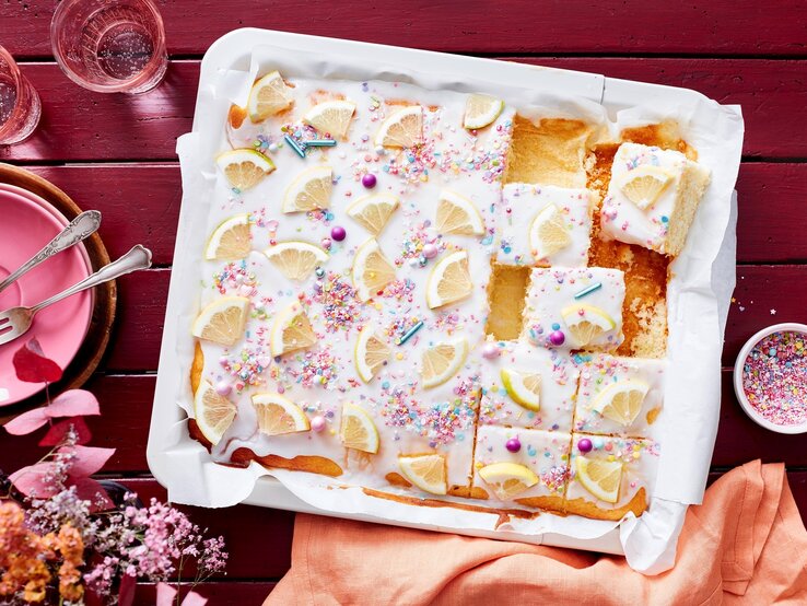 Auf einem weißen Teller liegt ein saftiger Zitronen-Buttermilchkuchen. Er ist mit Puderzuckerglasur und Zuckerstreuseln dekoriert und mit frischen Zitronen bedeckt. 