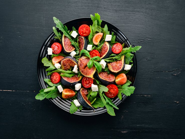 Vogelperspektive: Ein Feigen-Salat mit Rucola und Feta auf einem dunklen Teller, der auf einem dunken Untergrund steht.