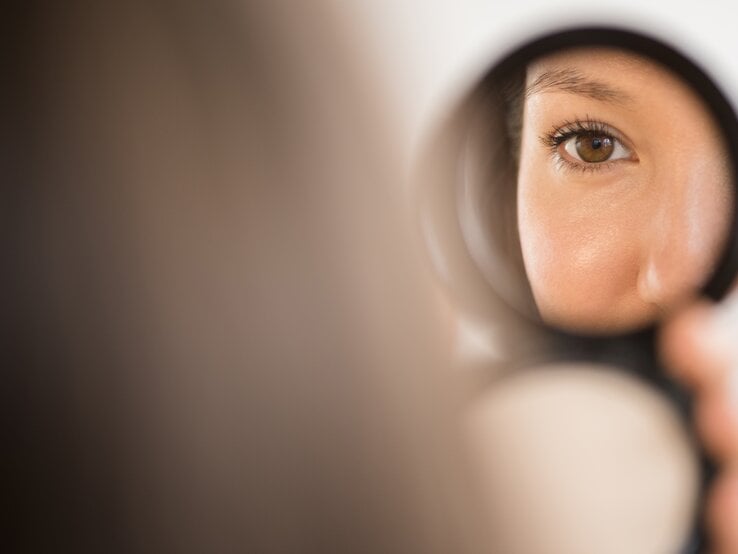 Eine Frau schaut in einen kleinen Handspiegel – nur ihr Auge und ihre Nase sind zu sehen.