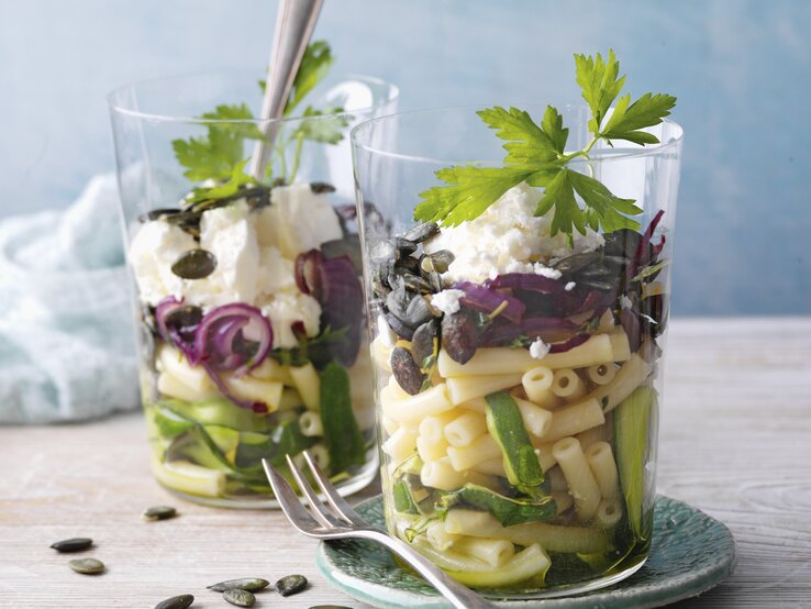 Zucchini-Makkaronisalat mit Feta im Glas. Daneben eine Gabel und im Hintergrund ein Küchentuch. 