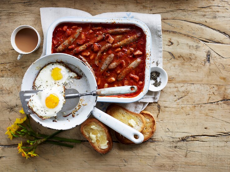 Wurstauflauf mit Bohnen und Speck in Tomatensoße, serviert mit krossen Spiegeleiern und frisch getoasteten Brioche-Scheiben. 