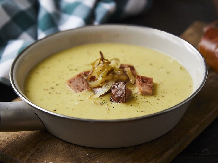 Auf einem Tisch steht eine Pfanne mit einer Kartoffelsuppe mit gebratenem Leberkäse. Sie sieht würzig und aromatisch aus. 