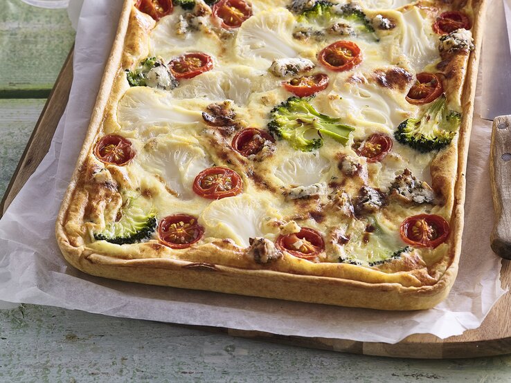 Zu sehen ist ein Blechkuchen mit Tomaten, Blumenkohl, Brokkoli, Wurst und überbackenem Käse in der Draufsicht auf Backpapier und Holzbrett. Rechts Messer mit Holzgriff. Alles liet auf grün-braunem Tisch mit einzelnen Brettern.  