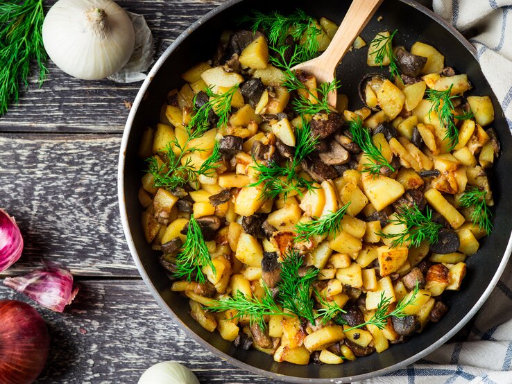 Draufsicht: Eine Pfanne mit Bratkartoffel mit Birne und Pilzen und Dill steht auf Holz. Naneben liegen eine weiße und eine rote Zwiebel sowie frischer Dill.