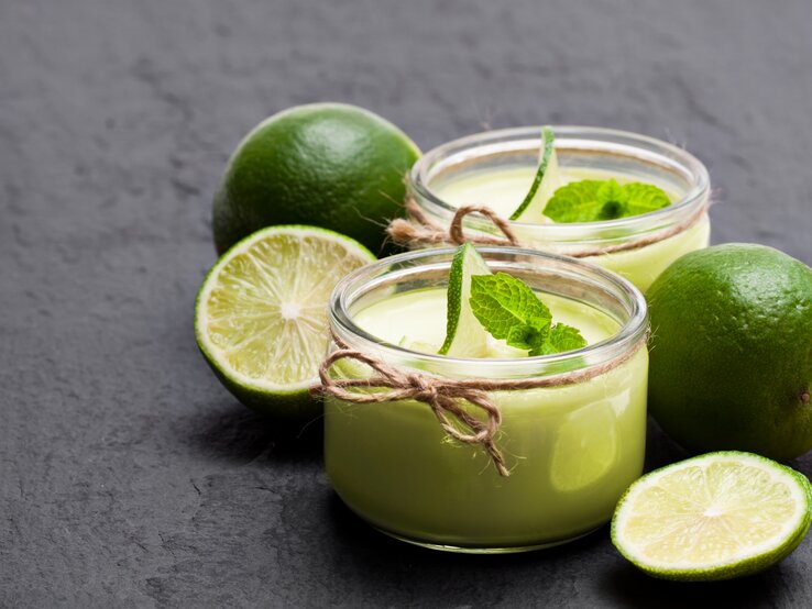 Frontalsicht: Auf einem Schiefer-Untergrund stehen zwei Gläser Limetten-Joghurt mit Minze. Drum herum liegen zwei ganze Limetten und zwei Limettenhälften.