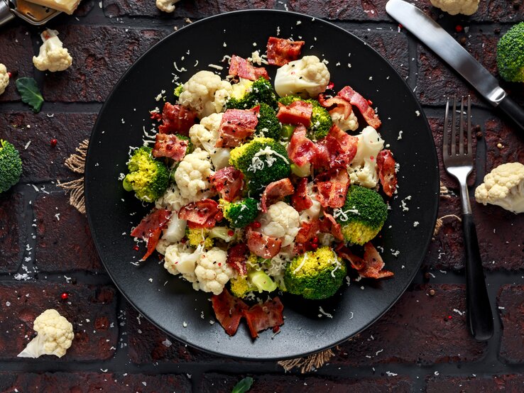 Ein Teller Blumenkohl-Brokkoli-Salat mit Parmaschinken, Parmesan und Sesam, drumherum liegen Kohlröschen und Besteck.