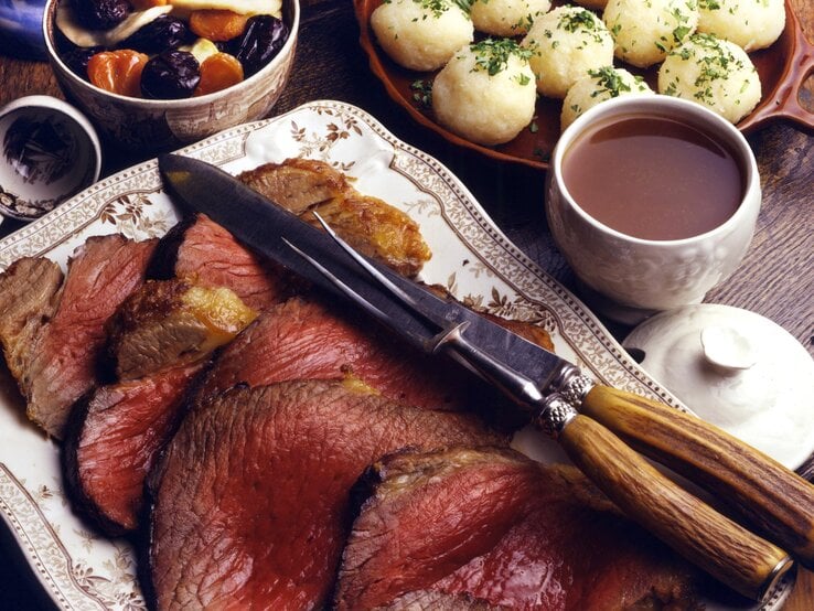 Angeschnittener Sauerbraten auf einem weißen Servierteller, daneben steht eine schale voll Klöße.