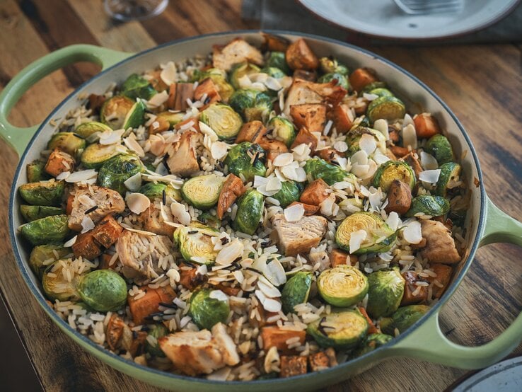 Eine Rosenkohl-Huhn-Reis-Pfanne in einer großen, grünen Pfanne mit hohem Rand. Die Reispfanne ist angerichtet mit Mandelblättchen und steht auf einem braunen Holztisch.