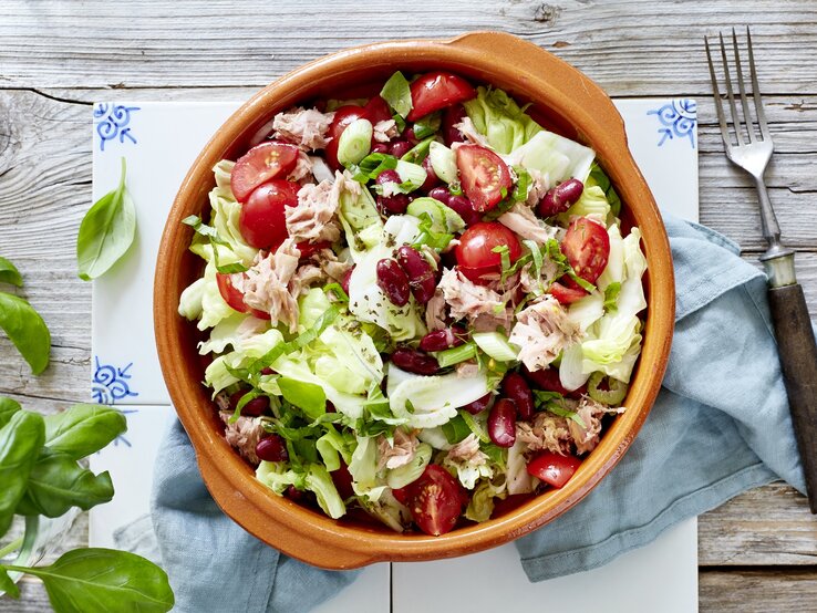 Bunter Thunfisch-Salat mit Kidneybohnen, Cocktailtomaten, Kräutern und Eisbergsalat, serviert in einer rustikalen Tonschale auf einem Holztisch, von oben fotografiert. 