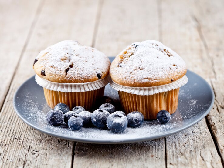 Frontalsicht: Auf einem Holzuntergrund steht ein grauer Teller mit zwei Joghurt-Muffins mit Beeren. Neben den Muffins liegen frische Blaubeeren.