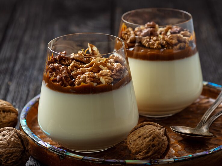 Weihnachtliches Panna Cotta mit Gewürzkaramell und Walnüssen im konischen Glas auf Holzteller mit Perlmuttrand auf dunklem Holztisch in der Frontalansicht. Ganze Walnüsse und zweit Silberlöffel als Dekoration.