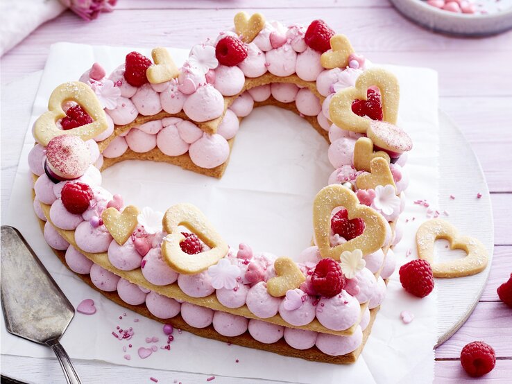 Auf einem Teller liegt ein Herz-Lettercake. Er ist dekoriert mit Zuckerperlen, Keks-Herzen, Himbeeren und rosa Creme. 