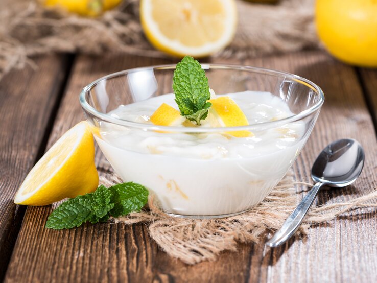 Frontalsicht: Auf einem Holztisch steht eine kleine Schüssel aus Glas mit einer Buttermilch-Creme mit Zitrone. Die ist mit Minze und Zitronenscheiben garniert. Drum herum liegen Zitronen, Minzblätter und ein kleiner Löffel.