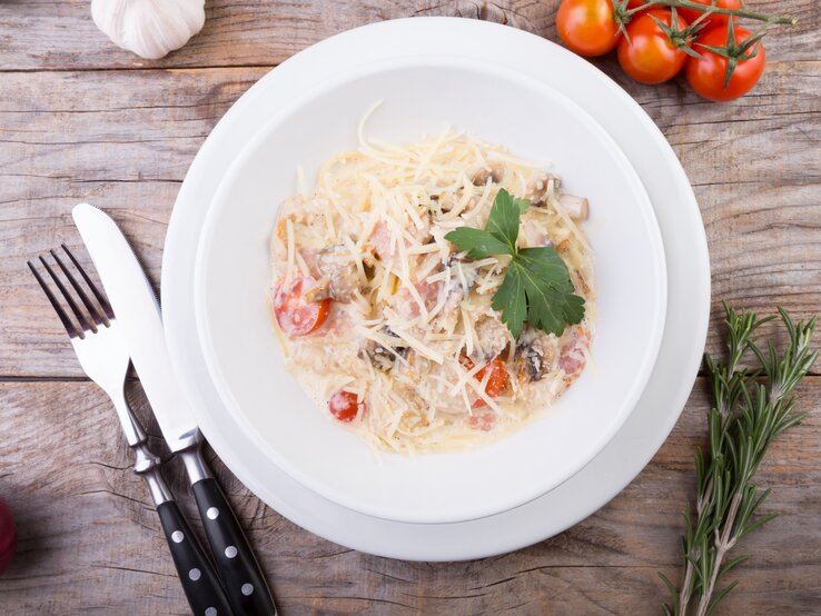 Weißer Teller belegt mit Spaghetti mit Tomaten, Pilzen und Speck in Mascarponesoße auf hölzernem Untergrund. 