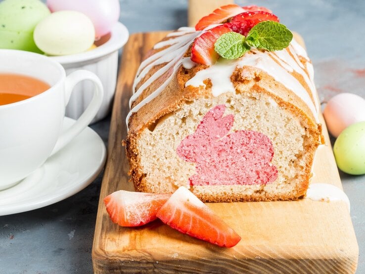 Auf einem Holzbrett liegt ein Osterhasenkuchen aus der Kastenform. Man erkennt einen rosa Hasen im Teig. Der Kuchen ist mit Zuckerguss überzogen. 