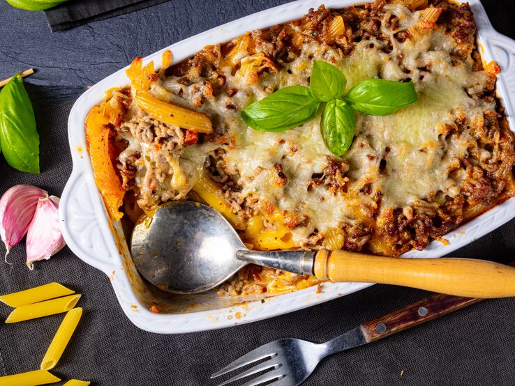 Auflauf mit Penne und Bolognese in einer Foorm, angerichtet auf dunklem Untergrund – umgeben von Basilikum, Knoblauch und Besteck.