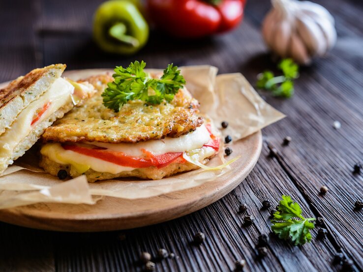 Die Low-Carb-Panini mit Mozzarella und Tomate auf einem Brett. Dahinter verschiedene Zutaten auf einem hölzernen Untergrund.