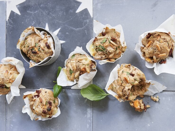 Vegetarische Gemüse-Pizza-Muffins mit Pinienkernen und frischem Basilikum, eingewickelt in Backpapier und drappiert auf einem hellen Metalltisch. 