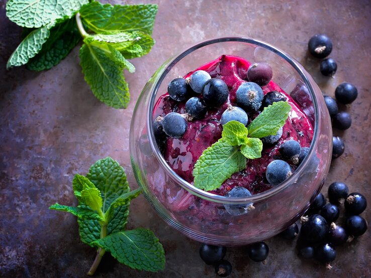 Draufsicht: Auf einem dunklem Untergrund steht ein Dessertglas mit einem Johannisbeer-Sorbet mit Prosecco. Daneben liegen schwarze Johannisbeeren sowie ein paar Blätter Minze.