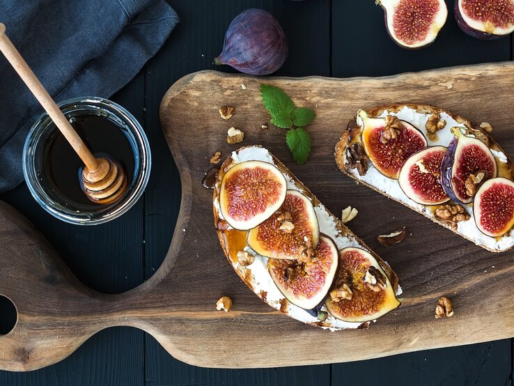 Zwei Brotscheiben mit Ricotta, Feigenund Honig auf einem dunklen Tisch. Daneben einige frische Feigen, ein Glas Honig und ein blaues Tuch.