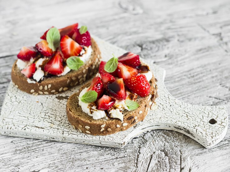 Draufsicht: zwei Erdbeer-Sandwiches mit Ricotta und Minze liegen auf einem hellen Holzbrett, das auf einem hellem Untergrund aus Holz liegt.