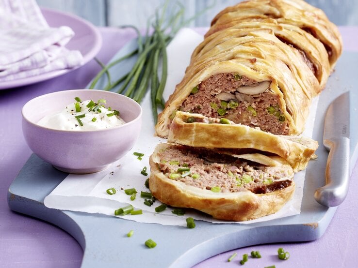 Hackbraten im Blätterteigmantel mit einer Füllung aus Hackfleisch, Pilzen, Erbsen und Schinken. Angeschnitten serviert auf einem Holzbrett und serviert mit frischem Schnittlauchschmand. 