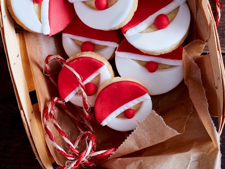 Nikolaus-Kekse auf Krepp in Bastkorb auf dunklem Holztisch aus der Vogelperspektive. Rotes und weißes Fondant mit roter Fondantkugel als Nase ergeben die Gesichter. Rot-weiße Schnur als Deko rechts neben dem Korb und auf dem Korb. 