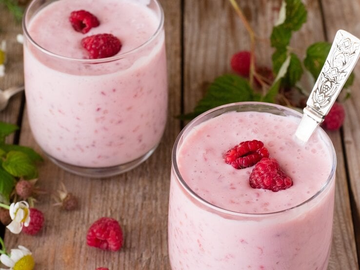 Zwei Gläser mit dem Kokos-Himbeer-Joghurt auf einem rustikalen Holztisch mit frischen Himbeerzweigen.