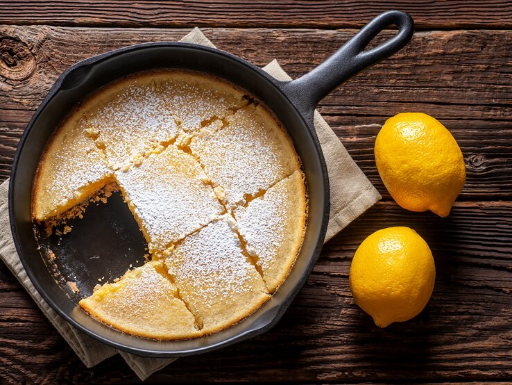 Zitronenkuchen ohne Backen aus der Pfanne in schwarzer Gusspfanne auf beigem Tuch und dunklem Holz. Daneben zwei Zitronen. Aufnahme aus der Vogelperspektive.