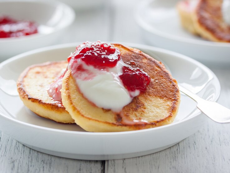 Hüttenkäse Pfannkuchen auf weißem Teller mit Joghurt und Himbeere verfeinert. Drei Portionen auf einem hellem Holztisch.