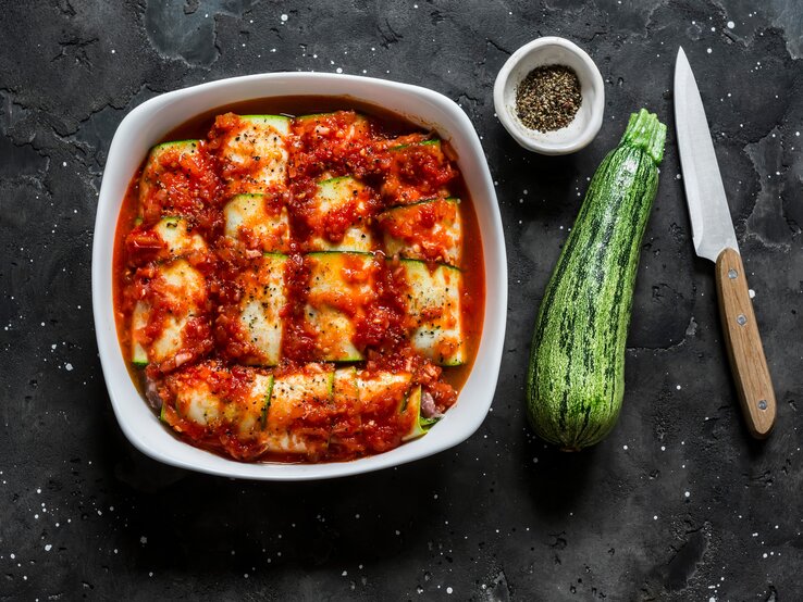 Low Carb Zucchini-Cannelloni mit Spinat-Ricotta-Füllung und fruchtiger Tomatensoße in Auflaufform auf schwarzem Untergrund. Rechts kleine Schale mit buntem Pfeffer. Daneben eine Zucchini und ein Messer mit Holzgriff. Aufnahme aus der Vogelperspektive. 