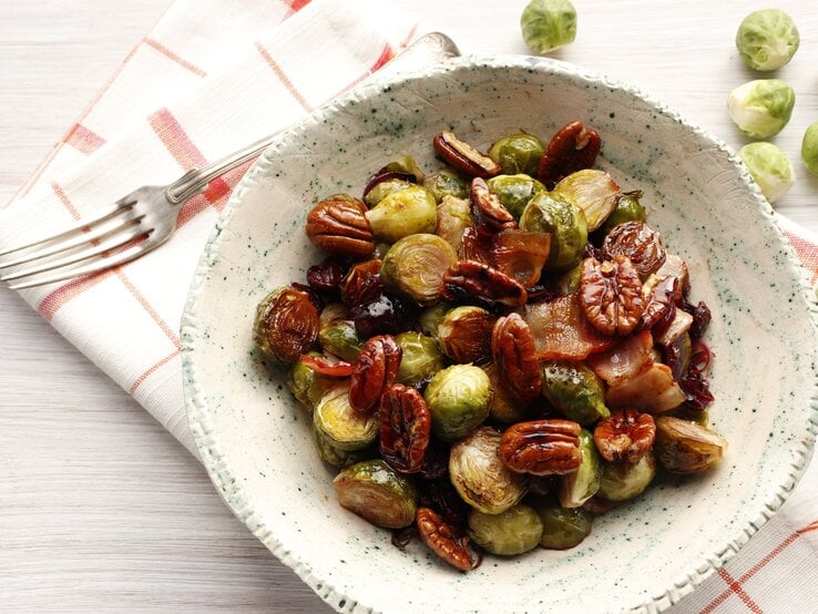 Ein Rosenkohl-Salat mit Nüssen und getrockneten Cranberries in einer weißen, gemusterten Schale steht auf einem karierten Geschirrtuch auf einem weißen Tisch. Neben der Schale liegt eine Gabel und einige einzelne Rosenkohlröschen.