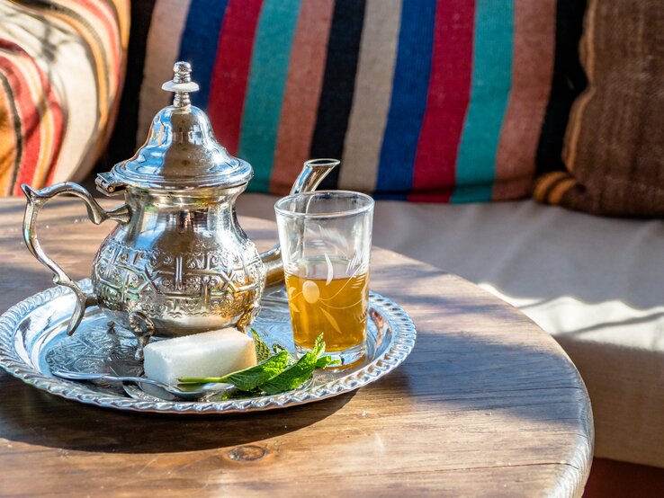 Auf einem metallenen Tablett stehen eine marokkanische Teekanne neben einem mit Minz-Tee gefüllten Teeglas, einem Stück Zucker und einem Minz-Blatt zur Garnierung.  | © GettyImages/Craig Hastings